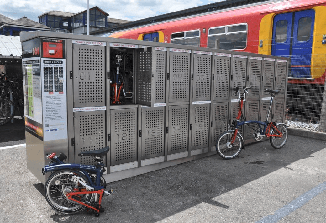 Casilleros de almacenamiento para bicicletas plegables en una estación de tren con bicicletas plegables estacionadas y un tren colorido en el fondo