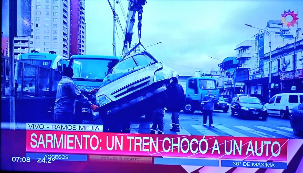 Noticia en televisión que muestra un accidente en el que un tren Sarmiento chocó a un auto en Ramos Mejía. Imagen de un automóvil siendo retirado con una grúa en la calle, rodeado de personas y vehículos.