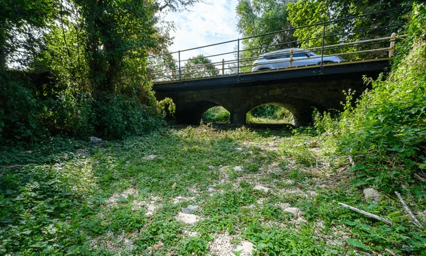 Puente de piedra bajo el cual el lecho de un río está completamente seco, cubierto de vegetación y piedras visibles. Al fondo, un coche estacionado en el puente y árboles frondosos rodean la escena, sugiriendo sequía o disminución drástica del caudal del río.