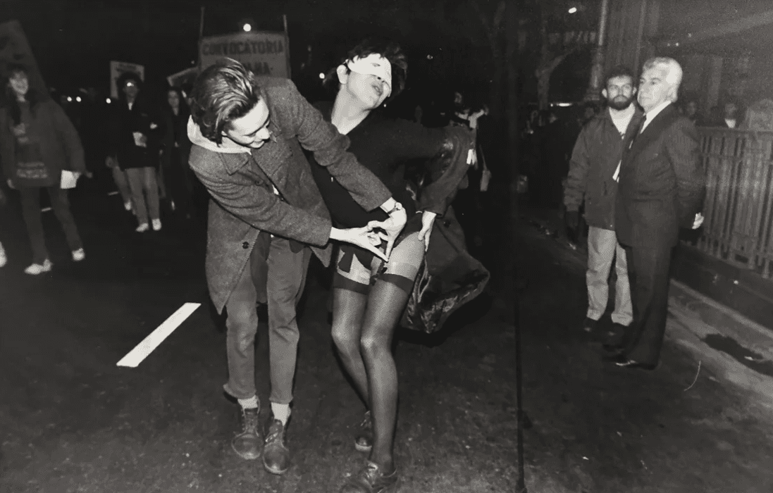 Fotografía en blanco y negro de una marcha del orgullo, una persona vestida con medias de red y máscara interactúa de manera provocativa con otra persona, mientras varios observadores miran la escena, el ambiente parece ser nocturno, con pancartas de fondo, descripción adicional menciona la evolución de las marchas del orgullo en Argentina, el recorrido histórico desde Plaza de Mayo hasta el Congreso, y el contexto de politización de la comunidad LGBT+ después de 2001, escenarios nombrados en honor a activistas Carlos Jáuregui y Nadia Echazú.