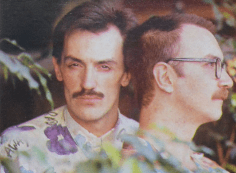 Fotografía de dos hombres, uno mirando hacia la cámara con expresión seria y bigote, mientras el otro está de perfil mirando hacia la derecha, ambos lucen camisas con patrones de colores, fondo con vegetación, la imagen transmite un estilo íntimo y reflexivo,  relacionada con temas de activismo o derechos LGBTQ+.