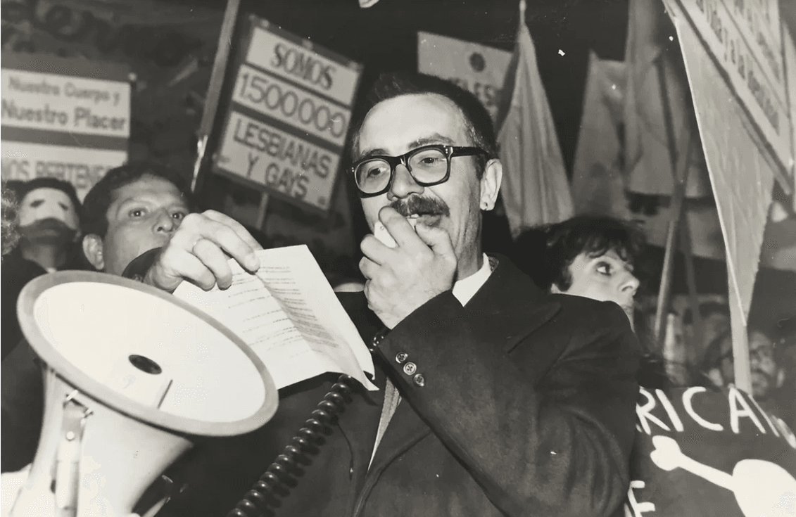 Fotografía en blanco y negro de una manifestación, en el centro aparece un hombre de bigote y gafas, hablando a través de un megáfono y sosteniendo una hoja de papel, detrás de él se ven personas con pancartas que incluyen mensajes como "Somos 1.500.000 lesbianas y gays" y "Nuestro cuerpo y nuestro placer", la escena transmite un ambiente de lucha y activismo en favor de los derechos de la comunidad LGBTQ+.