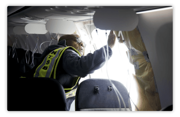 La imagen muestra a un inspector o investigador de la NTSB (National Transportation Safety Board) dentro de un avión. La persona está usando un chaleco de alta visibilidad con las siglas "NTSB" en la espalda y parece estar examinando o inspeccionando la estructura de la ventana del avión. Se observan cables y partes del avión expuestas, lo cual sugiere que la aeronave está siendo investigada o revisada posiblemente tras un incidente o por mantenimiento detallado. La iluminación de la escena está dirigida hacia el área de inspección, enfocando la actividad del investigador.