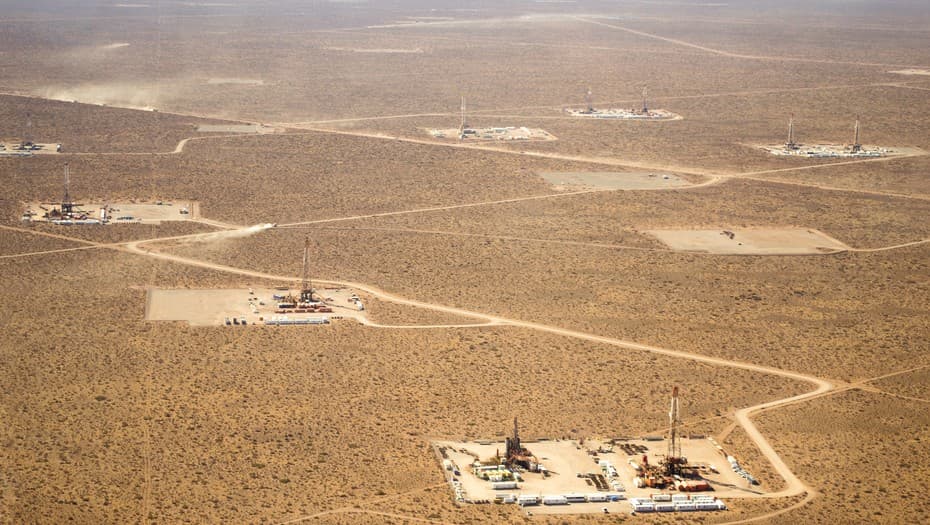 Fotografía aérea de un área de explotación petrolera en una zona árida y extensa, se observan múltiples plataformas de perforación distribuidas en la tierra, conectadas por caminos de tierra que serpentean entre las instalaciones, el paisaje está mayormente despoblado y cubierto de vegetación baja típica de zonas desérticas, la imagen destaca la extensión de la operación y el impacto en el entorno natural, representando un espacio dedicado a la extracción de recursos en una región remota.